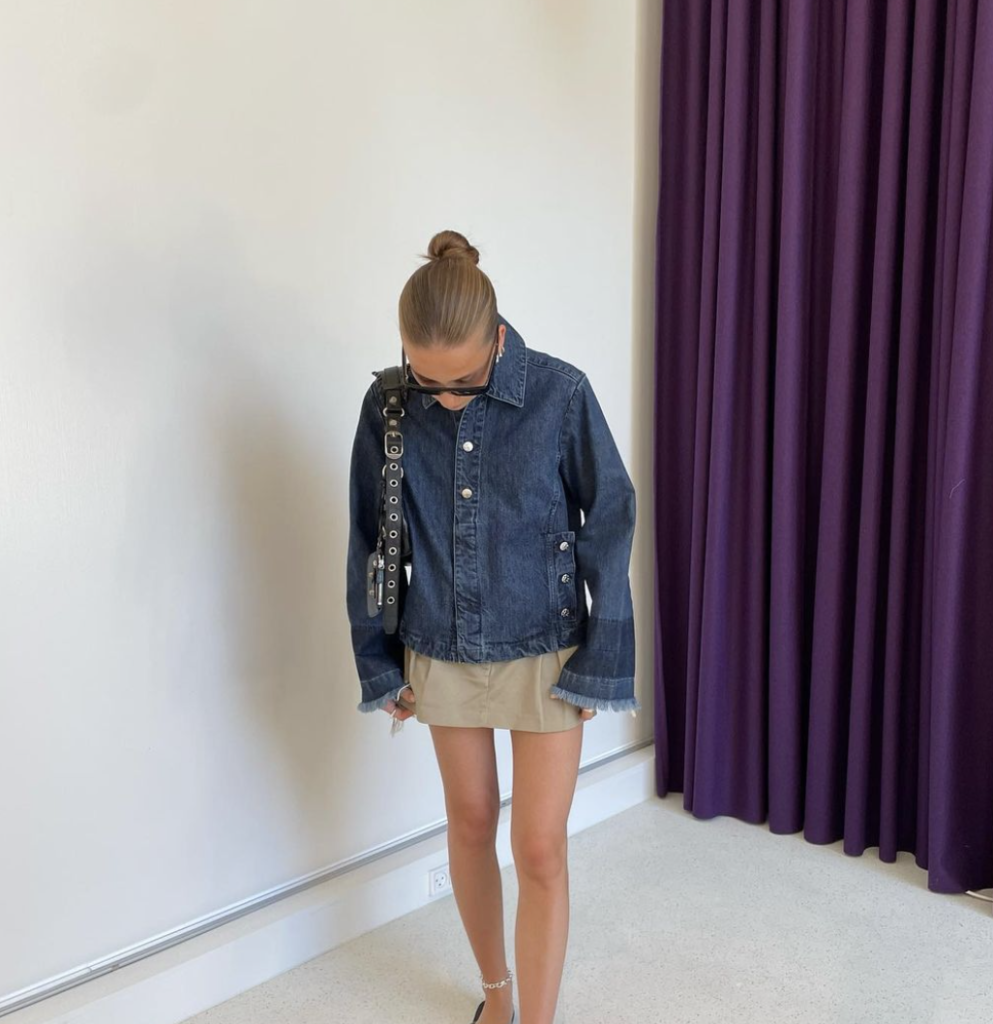 Person in a unique denim jacket and a khaki skirt, standing in a room with a white wall and purple curtains.